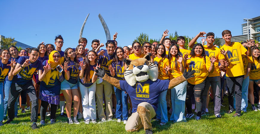 Students in blue and gold surrounding Rufus as they gather in front of the Beginnings Sculpture.