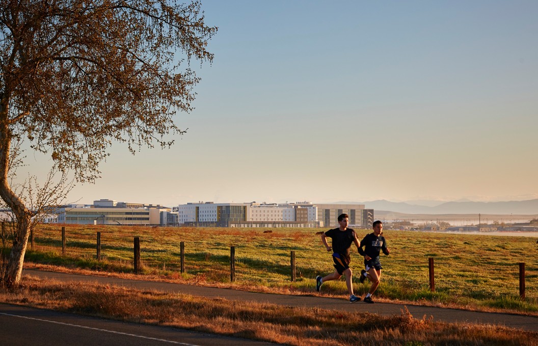Two students running on a trail along Old Lake Road, with buildings from UC Merced pictured on the background. against a sunrise.
